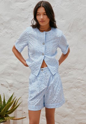 Femme portant une chemise à manches courtes à carreaux bleu clair et blanc, boutonnée, et un short assorti, debout contre un mur blanc texturé.