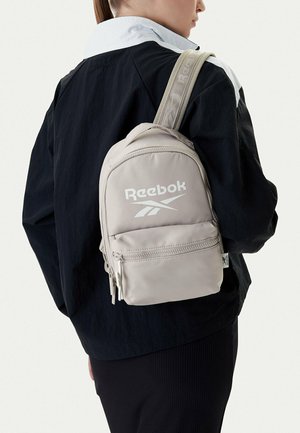 Persona vistiendo una mochila Reebok gris claro sobre un hombro, con una chaqueta blanca y negra, vista desde atrás y de lado.