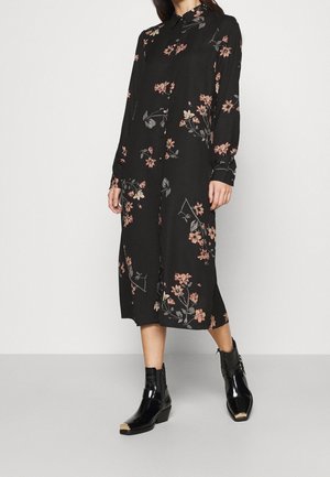 Kvinde iført en sort midi skjortekjole med pink blomstermønstre og sorte ankelstøvler med metallic forkanter, stående på en grå gulv.