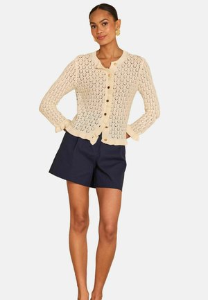 Vrouw draagt een beige gebreid vest met ruchtrandjes en donkerblauwe korte broek, staat met handen in de zij tegen een effen achtergrond.