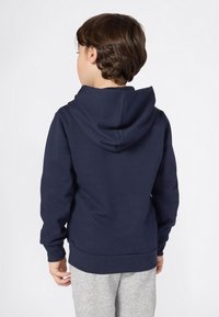 Kind met kort bruin haar, gekleed in een donkerblauwe hoodie en lichtgrijze trainingsbroek, met de rug naar de camera gekeerd tegen een effen witte achtergrond.