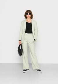 Femeie îmbrăcată într-un costum verde deschis cu bluză neagră, ochelari de soare negri, ținând o geantă neagră matlasată și adidași alb-negru, stând în fața unui perete alb.