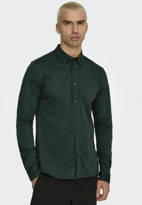 Camisa de manga larga, verde oscuro, de una tela suave con cuello tipo button-down, que presenta una tapeta frontal con botones negros y un dobladillo curvo.