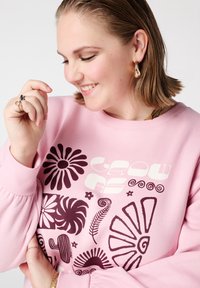 Lichtroze sweatshirt met gedurfde, donkere ontwerpen van bloemen, spiralen en een grafische tekst "GROW AS." Ronde halslijn met geribbelde manchetten.