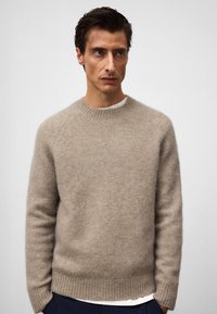 Beżowy sweter wykonany z miękkiego materiału, z okrągłym dekoltem, ściągaczami na mankietach i u dołu, oraz lekko luźnym krojem z widoczną teksturą.