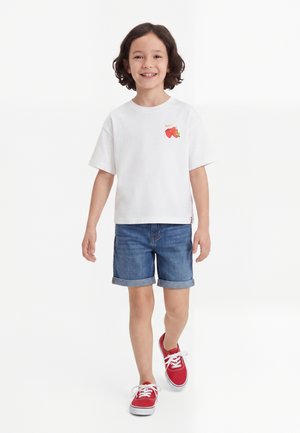 Niño sonriente con cabello rizado caminando, llevando una camiseta blanca con diseño de fresas, pantalones cortos vaqueros y zapatillas rojas con cordones blancos.