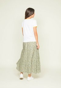 T-shirt blanc avec des bordures jaunes, associé à une longue jupe fluide à motifs floraux en vert et jaune. Le modèle porte des baskets blanches.