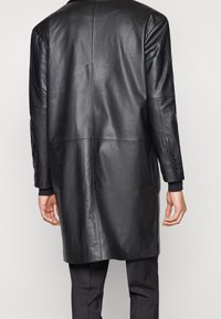 Mężczyzna w czarnym skórzanym długim płaszczu i czarnych spodniach, pokazany z tyłu na jednolitym jasnym tle.