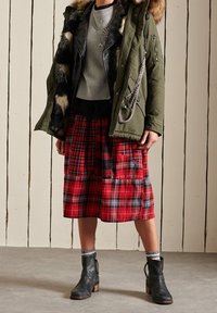 Olive green parka with fur trim, black leather jacket, gray sweater, and red plaid skirt. Black ankle boots with heel and gray socks visible.