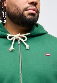 Κοντινό πλάνο ενός άνδρα με γένια που φοράει πράσινο φούτερ με φερμουάρ, λευκά κορδόνια και μικρό κόκκινο λογότυπο Levi’s στο στήθος.