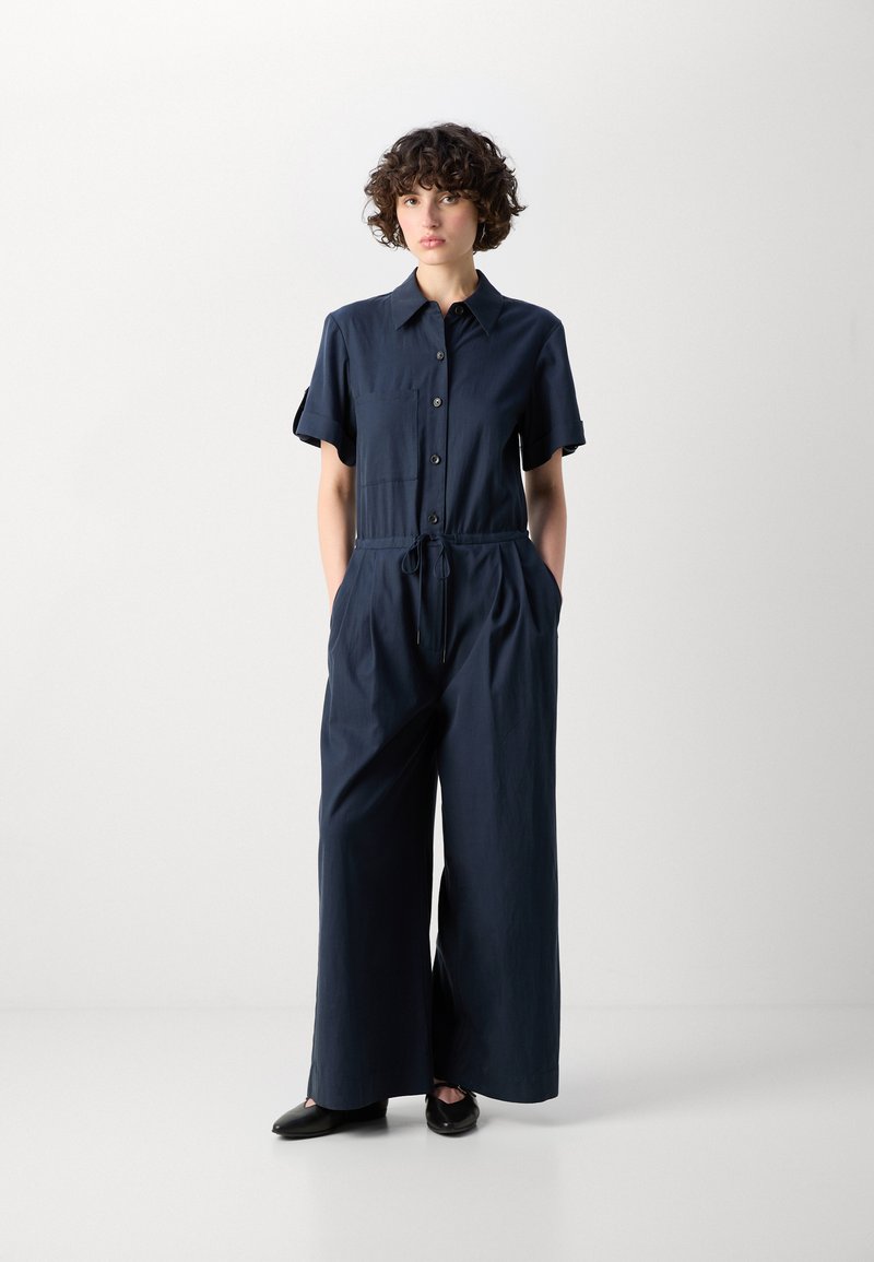 Navyblauer Overall mit kurzen Ärmeln, Knopfverschluss und Schnürbund. Verfügt über eine Vordertasche und ein weit geschnittenes Design. Glatte Stoffoberfläche.