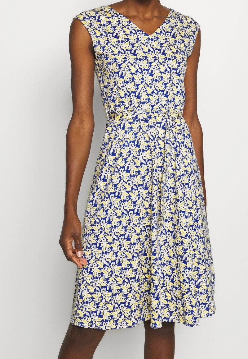 Robe sans manches longueur genou avec décolleté en V, taille cintrée, en tissu bleu à motif de petites fleurs jaunes et blanches.