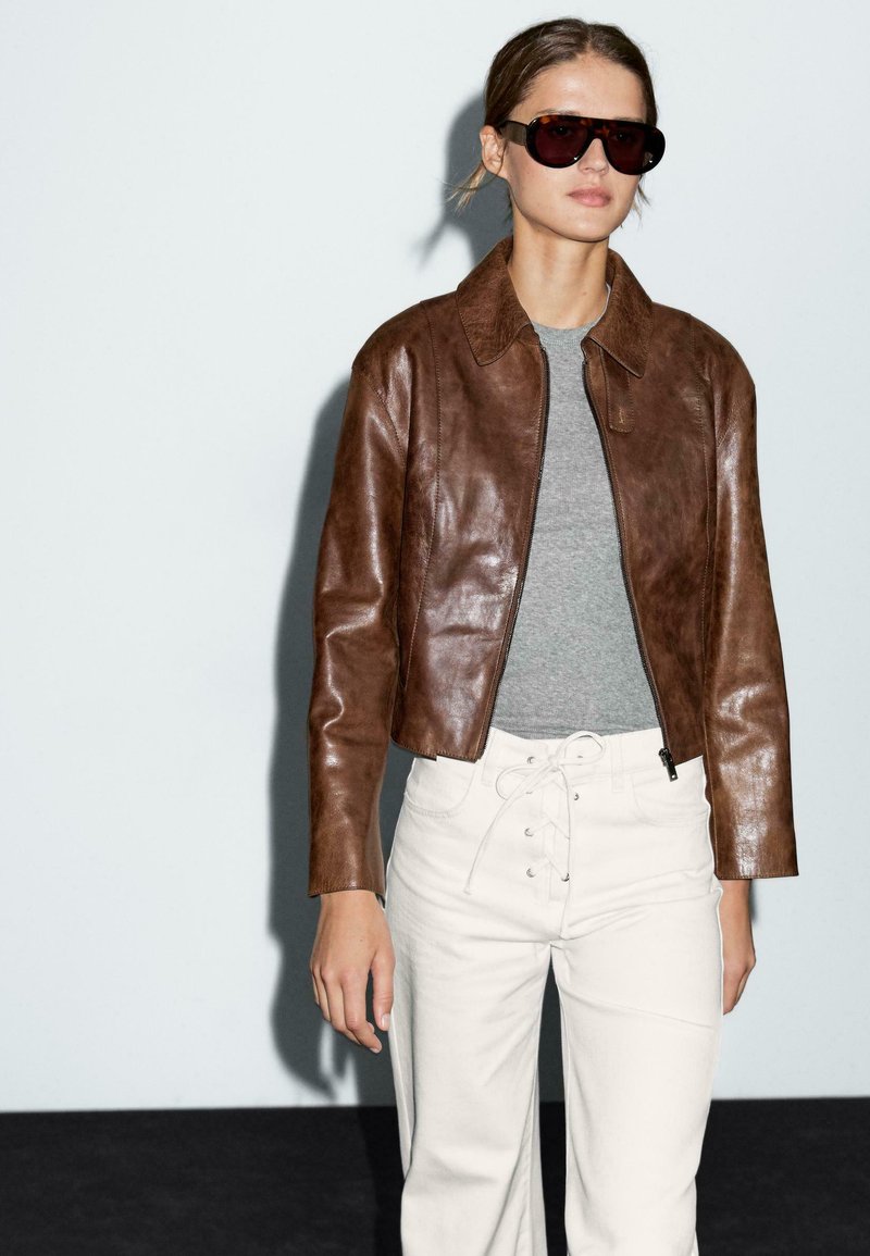 Veste cropped en cuir marron avec un col, assortie à un haut gris et un pantalon blanc à jambes larges. Texture lisse, sans motifs visibles.