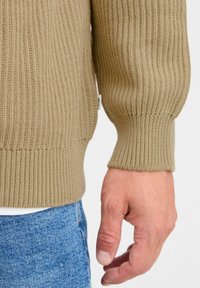 Beige ribbed sweater with a textured finish. Close-up of the cuff, featuring a fitted hem. Hand visible with blue denim fabric in the background.