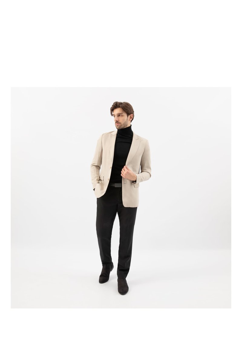 Man draagt een beige blazer, zwarte coltrui, zwarte broek en zwarte schoenen, staand tegen een effen witte achtergrond.