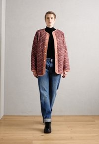 Rdeča jakna s vzorcem in kovinskimi gumbi, nošena čez črn turtleneck, v kombinaciji z modrimi kavbojkami in črnimi čevlji, na lesenem tleh.