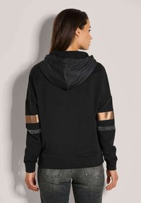 Schwarzer Hoodie mit einer glänzenden schwarzen Kapuze, goldenen und silbernen Akzenten an den Ärmeln und einem entspannten Schnitt. Weicher Stoff mit gerippten Bündchen und einem Bund.