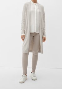 Lång beige cardigan med öppen framsida och fickor, lager på lager över en vit skjorta med krage, ihop med ljusbeiga byxor och vita sneakers.