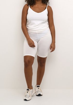 Femme debout portant un débardeur blanc, un short de vélo blanc et des baskets blanches sur fond clair.