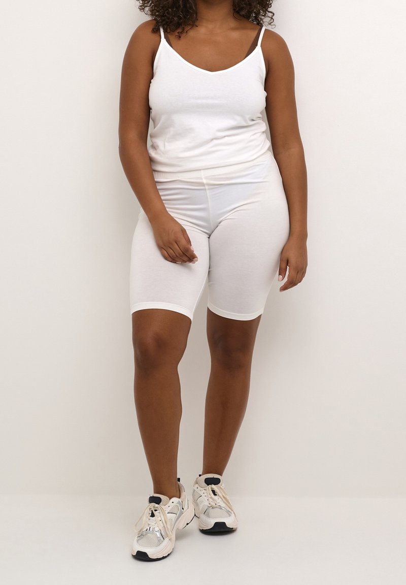 Femme debout portant un débardeur blanc, un short de vélo blanc et des baskets blanches sur fond clair.
