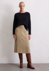 Pull en maille côtelée navy, jupe beige à motif floral bleu, et bottes en cuir marron jusqu'aux genoux, sur fond léger texturé.