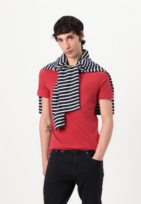 Rotes Kurzarm-T-Shirt mit kleinem Logo, kombiniert mit einem schwarz-weißen gestreiften Pullover, der über die Schultern getragen wird, und schwarzen Jeans.