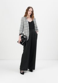 Šedý kostkovaný blazer přes černý top, kombinovaný s černými kalhotami se širokými nohavicemi a vysokým pasem, a špičatými černými botami, držící černou tašku.