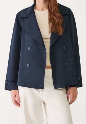 Woman wearing a navy blue double-breasted jacket over a beige top and white pants, standing against a plain light background.