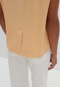 Perzikkleurige mouwloze top met een rechte snit en een split aan de achterkant, gecombineerd met witte jeans met zakdetails. Zachte, gladde textuur.