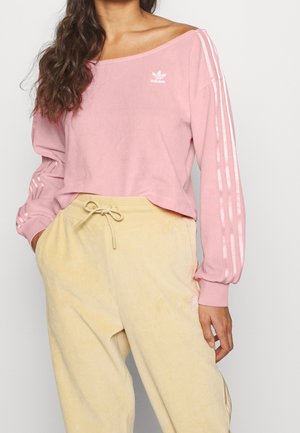 Kobieta ubrana w różową bluzkę Adidas z długim rękawem i paskami na rękawach oraz bladożółte spodnie ze ściągaczem, ręce w kieszeniach.