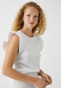 Witte katoenen t-shirt met gerimpelde schouders en een ronde halslijn, gecombineerd met een gestructureerde, lichtgekleurde rok. Model heeft krullend haar.