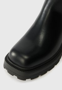 Crne kožne čizme s zaobljenim nosom, teksturiranim potplatom s utorima i vidljivim šavovima duž rubova. Glatka površina gornjeg materijala.