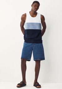 Sandali neri, pantaloncini di jeans e una canottiera con una parte superiore bianca, una sezione centrale a righe blu e una parte inferiore blu scuro.