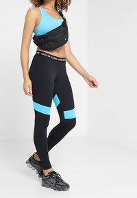 Mustat legginssit sinisillä yksityiskohdilla, yhdistettyinä siniseen toppiin ja mustaan ristikotilaukseen. Suunniteltu suorituskykyä ja aktiivista käyttöä varten.