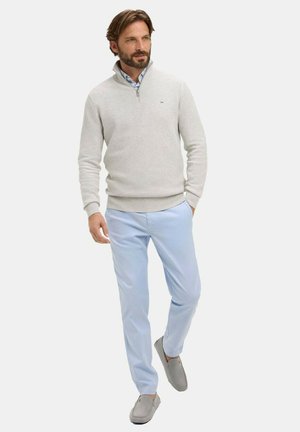 Homme barbu portant un pull gris clair à fermeture éclair quart de tour, une chemise bleue avec col, un pantalon bleu clair et des chaussures grises sans lacets, marchant les mains dans les poches.