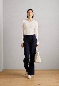 Hvit langermet topp, marineblå stripebukser, kremfarget veske og beige sko. Slim-fit design med myk tekstur, vist mot en nøytral bakgrunn.
