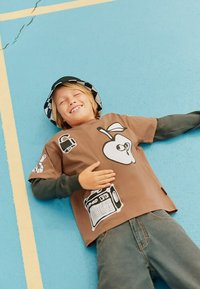 T-shirt marron à manches courtes avec des imprimés graphiques, dont une pomme et une radio. Porté avec une chemise verte à manches longues en dessous et un pantalon en denim.