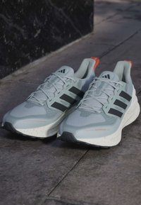 Et par grå Adidas løbesko med sorte striber og orange trækkende faner hvilende på en stenovertflade.