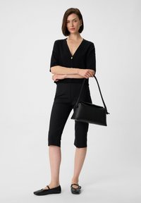 Schwarzes, kurzes Oberteil mit kurzen Ärmeln und V-Ausschnitt, kombiniert mit schwarzen Capri-Hosen. Das Model hält eine schwarze Handtasche mit einem langen Trageriemen.