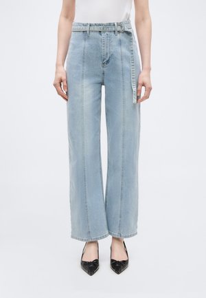 Nő világoskék, magas derekú, bőszárú farmert visel, amelyhez egy hozzáillő anyagból készült öv tartozik, fekete hegyes orrú magassarkú cipőben, egyszínű háttér előtt.