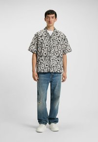 Camicia a maniche corte con un motivo grafico astratto in bianco e nero, abbinata a jeans blu strappati e sneaker bianche.