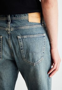 Světle modré džíny vyrobené z denimové tkaniny s koženou nášivkou označenou "JACK & JONES." Zadní kapsy s zakřivenými švy.