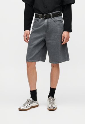 Άτομο που φορά μεσαίου μήκους γκρι τζιν σορτς, μαύρο πουκάμισο μακρυμάνικο, μαύρη ζώνη, μαύρες κάλτσες και λαμπερά ασημένια αθλητικά παπούτσια, όρθιο.