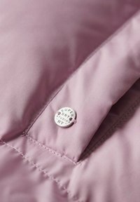 Rosa puffjacka i tyg med en slät textur, prydd med en metallisk rund logoknapp med "Super Dry Studios NY" ingraverat.