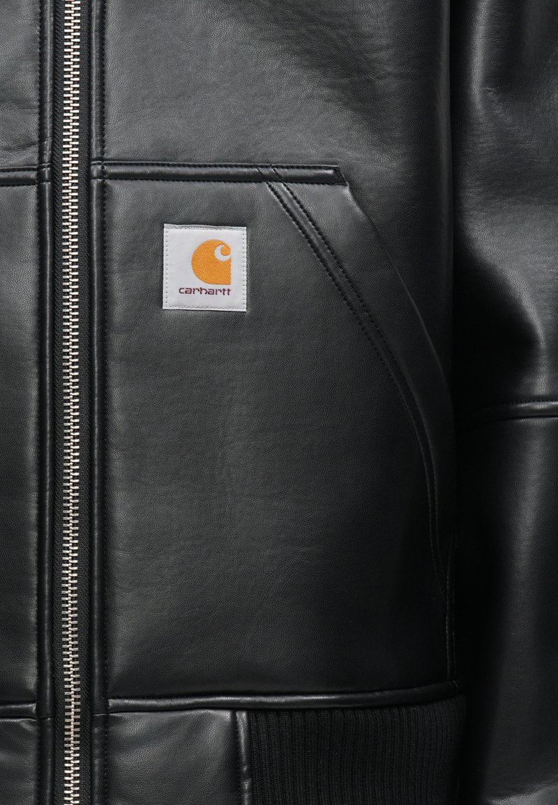 Schwarze Lederjacke mit einem Reißverschluss vorne, gerippten Bündchen und einem quadratischen weißen Etikett mit einem orangefarbenen Logo auf der linken Brust.