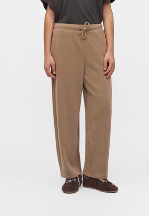 Personne portant un pantalon beige ample avec une taille à cordon et des chaussures marron foncé à lacets, debout devant un fond uni.