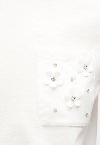 Άσπρη υφασμάτινη τσέπη διακοσμημένη με λευκές εφαρμογές λουλουδιών και μικρές διάφανες στρας σε ένα λευκό ένδυμα.