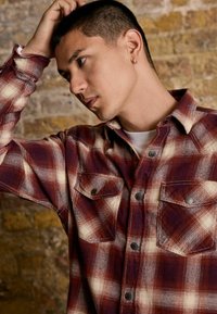 Young man with short hair and small hoop earring, wearing a red and cream plaid shirt, touching his head against a brick wall background.