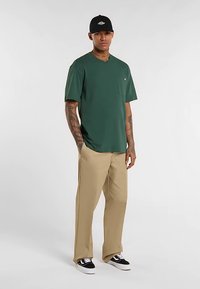 Camiseta verde de manga corta con bolsillo, pantalones beige, gorra negra y zapatos negros. Tela suave, corte relajado y diseño sencillo.