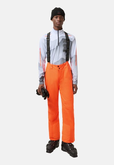 Jasnopomarańczowe spodnie narciarskie z regulowanymi szelkami, w zestawie z szarym longsleeve'em z motywem górskim i czarnymi rękawiczkami.